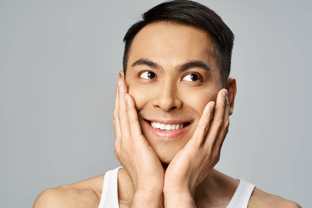 Bello uomo asiatico con un sorriso sereno, toccandosi delicatamente il viso in una rilassante routine di cura della pelle in uno studio grigio. - Foto, immagini