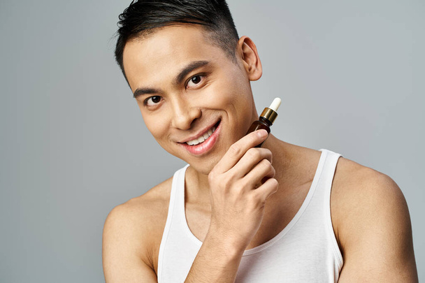 Porträt eines gut aussehenden asiatischen Mannes, der in einem grauen Studio eine Flasche Serum in der Hand hält, um seine Hautpflege zu verbessern. - Foto, Bild