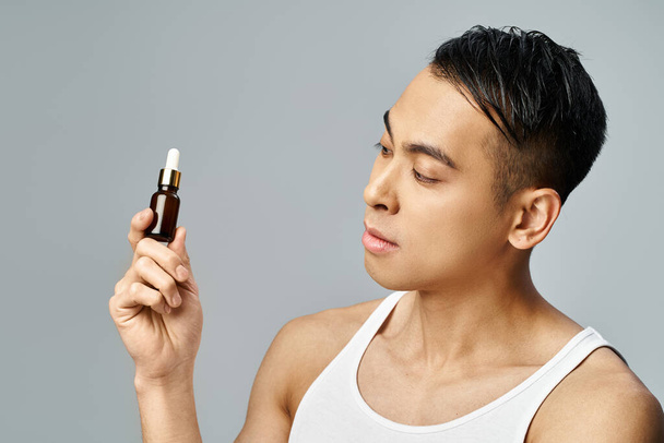 Un hombre asiático guapo en un estudio gris delicadamente sostiene una botella de aceites esenciales, que encarna la relajación y el autocuidado. - Foto, Imagen