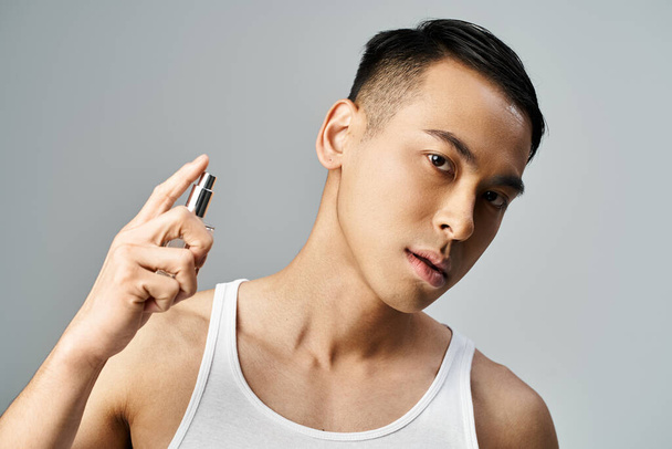 Un hombre asiático guapo en una camiseta sin mangas rociando su perfume de lujo en un estudio gris. - Foto, Imagen