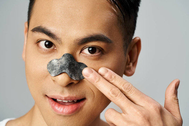 Ein gutaussehender asiatischer Mann in einem grauen Studio mit Schönheitsnasenpflaster, Hautpflege-Routine. - Foto, Bild