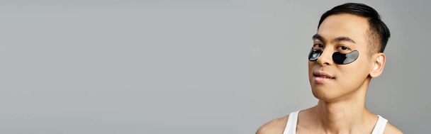 Портрет красивого азиата, раскачивающего повязку на глазу во время тренировки по уходу за кожей в серой студии. - Фото, изображение