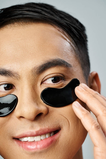 Красивий азіатський чоловік застосовує чорні плями очей для своєї шкіри рутини в сірій студії. - Фото, зображення