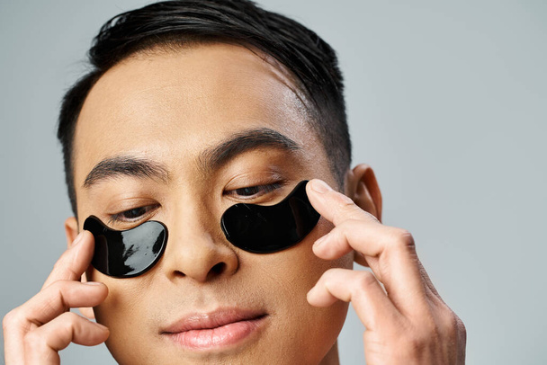 Un bell'uomo asiatico che tiene due cerchi neri fino agli occhi in uno studio grigio, concentrandosi sulla sua routine di bellezza e cura della pelle. - Foto, immagini