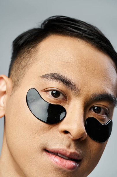Handsome Asian man with under eye patches during beauty and skincare routine in grey studio. - Photo, Image