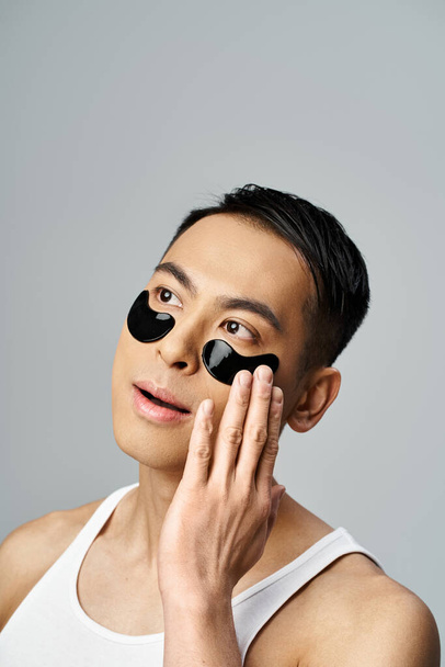 Hombre asiático guapo con manchas en los ojos negros, en una rutina de belleza y cuidado de la piel, en un entorno de estudio gris. - Foto, Imagen