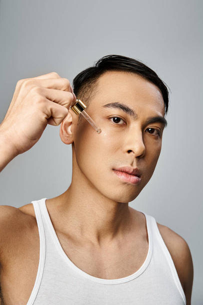 Een knappe Aziatische man met behulp van serum tijdens een beauty routine in een grijze studio setting. - Foto, afbeelding
