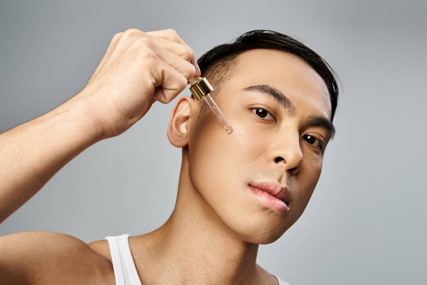 Un hombre asiático guapo sosteniendo pipeta con suero cerca de la mejilla durante una rutina de belleza y cuidado de la piel en un estudio gris. - Foto, imagen