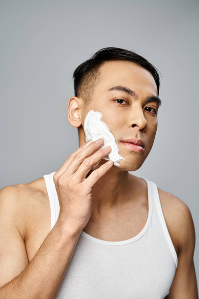 Schöner asiatischer Mann rasiert sich sanft sein Gesicht, die Augen fokussiert in einem grauen Studio-Setting. - Foto, Bild