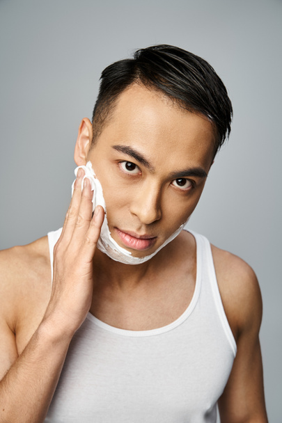 Handsome Asian man with shaving foam on face, gracefully shaving in a grey studio. - Photo, Image