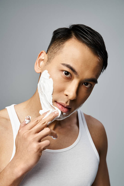 Um homem asiático bonito com espuma de barbear em seu rosto em um estúdio cinza, cuidadosamente barbear - Foto, Imagem