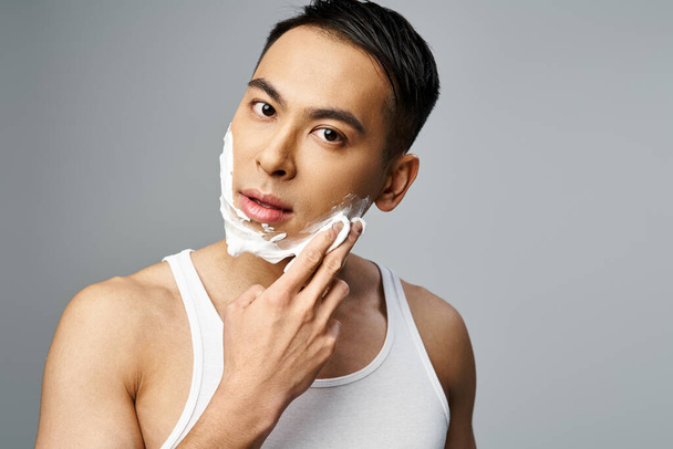 Красивий азіатський чоловік з піною для гоління на обличчі, ретельно голиться бритвою в сірій студії. - Фото, зображення
