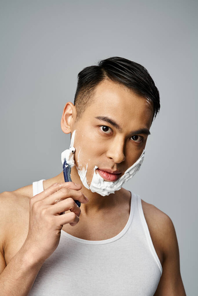 Un elegante hombre asiático vestido de negro se afeita la cara con una navaja de afeitar en un vibrante entorno de estudio rojo. - Foto, Imagen