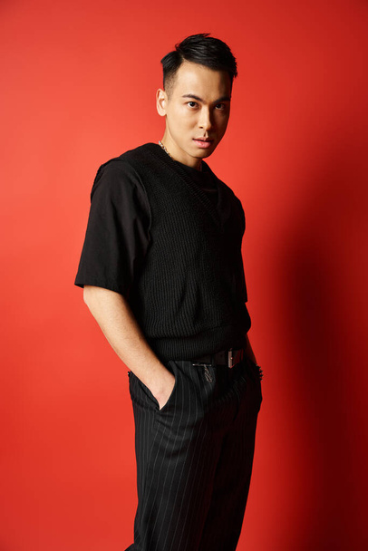 Um homem asiático elegante em trajes pretos confiantemente fica na frente de uma parede vermelha vibrante em um estúdio. - Foto, Imagem
