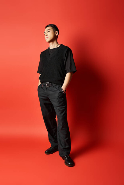 Un Asiatique à la mode en tenue noire se tient en confiance devant un mur rouge frappant dans un décor de studio. - Photo, image