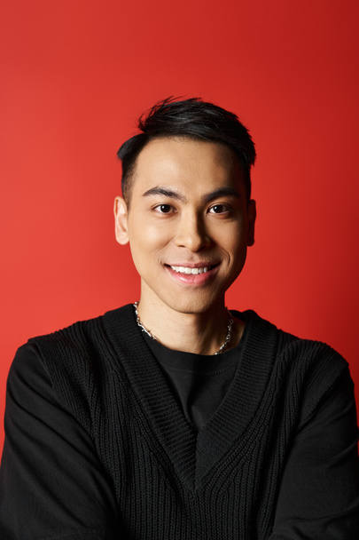 Un bel homme asiatique sourit chaleureusement à la caméra, portant un pull noir chic sur une toile de fond rouge vif studio. - Photo, image
