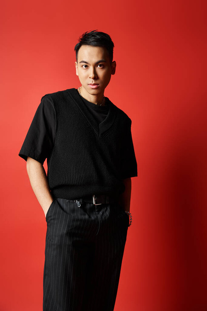 Ein stilvoller asiatischer Mann in schwarzer Kleidung steht selbstbewusst vor einer leuchtend roten Wand in einem Studio-Ambiente. - Foto, Bild