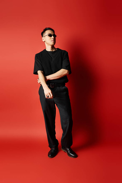 Ένας κομψός Ασιάτης, ντυμένος με μαύρη ενδυμασία, στέκεται με αυτοπεποίθηση μπροστά σε ένα τολμηρό κόκκινο φόντο σε ένα σκηνικό στούντιο. - Φωτογραφία, εικόνα