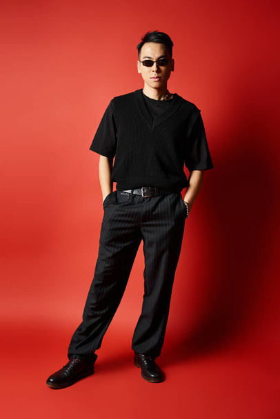 Tyylikäs aasialainen mies mustissa vaatteissa seisoo rohkeasti elinvoimaista punaista taustaa vasten silmiinpistävässä visuaalisessa koostumuksessa.. - Valokuva, kuva
