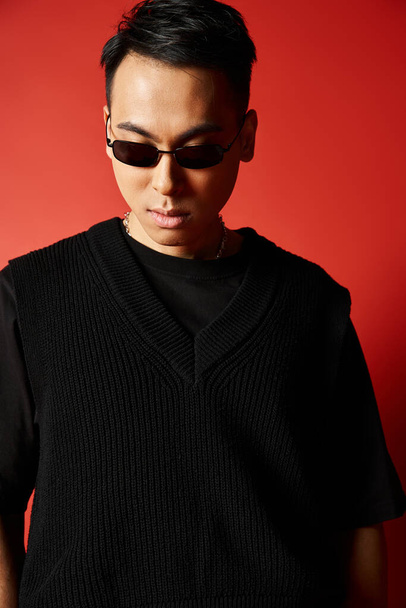 Een stijlvolle en knappe Aziatische man met een zonnebril en een zwarte trui tegen een levendige rode achtergrond. - Foto, afbeelding