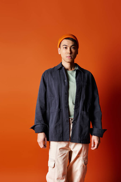 Yakışıklı bir Asyalı, şık giyinmiş bir şekilde stüdyoda canlı turuncu bir duvara karşı duruyor.. - Fotoğraf, Görsel