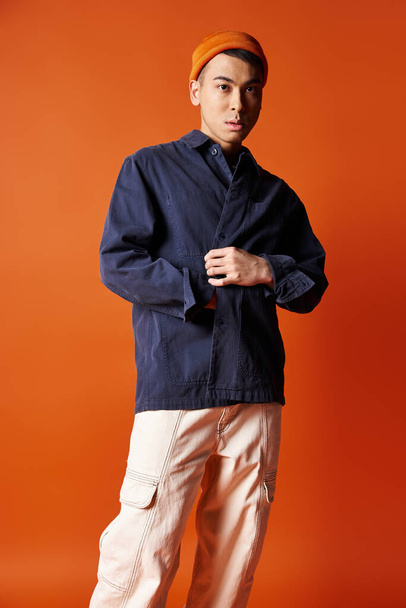 Красивый азиат в стильной одежде уверенно стоит на ярком оранжевом фоне в студии. - Фото, изображение