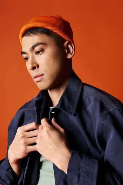 青いジャケットのハンサムなアジア人男性は,活気に満ちたスタジオの背景に対してオレンジ色の帽子に自信を持って立っています. - 写真・画像