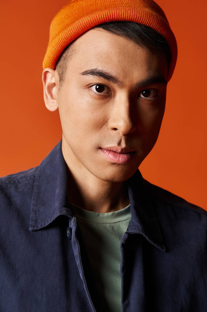 Красивый азиат в стильной синей рубашке и оранжевой шляпе уверенно стоит на оранжевом фоне в студии. - Фото, изображение