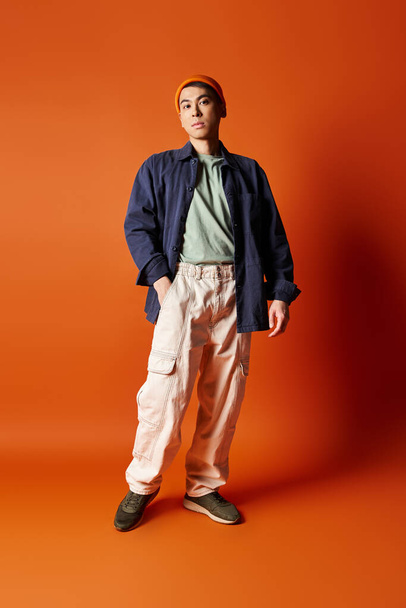 A handsome Asian man stands confidently in stylish attire against a vibrant orange background in a studio setting. - Photo, Image