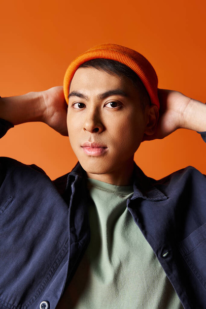 Hezký asijský muž v modré bundě a oranžovém klobouku, vyzařující styl a eleganci na pulzujícím oranžovém pozadí. - Fotografie, Obrázek