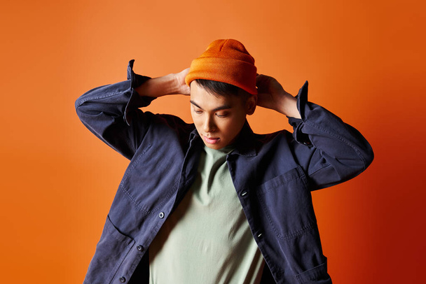 Un bell'uomo asiatico, vestito con abiti eleganti, indossa con sicurezza un cappello sulla testa su uno sfondo arancione.. - Foto, immagini