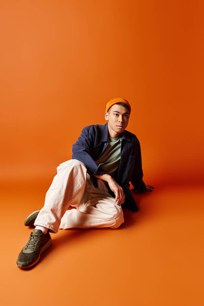 Красивий азіатський чоловік у стильному вбранні сидить на помаранчевому тлі, глибоко в думці або відображенні. - Фото, зображення
