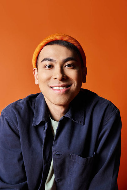 A handsome Asian man exudes style in a blue shirt and orange hat against a vibrant orange backdrop. - Photo, Image