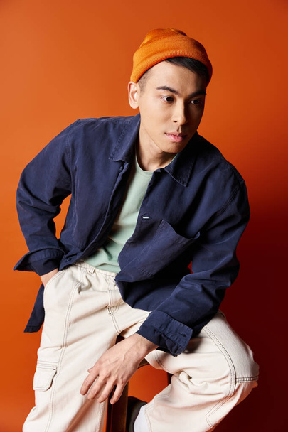 A handsome Asian man sits on a chair, styled in a blue jacket, against an orange background in a trendy studio. - Photo, Image