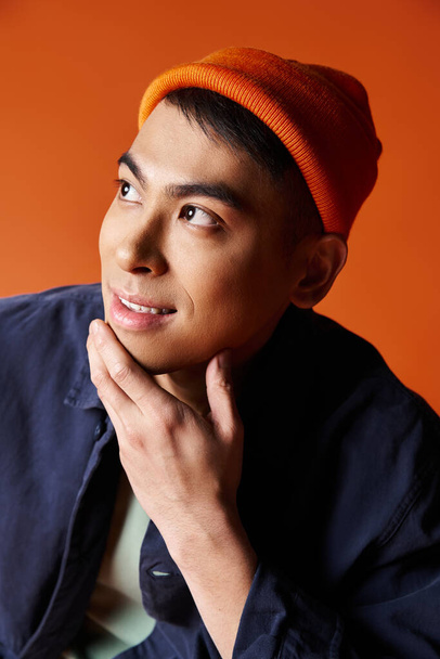 Een knappe Aziatische man draagt vol vertrouwen een blauw jasje en een oranje hoed tegen een levendige achtergrond. - Foto, afbeelding