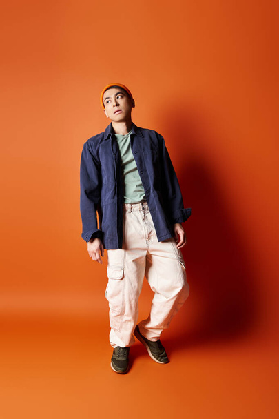 A handsome Asian man dressed in stylish attire stands confidently in front of a vivid orange wall in a studio setting. - Photo, Image