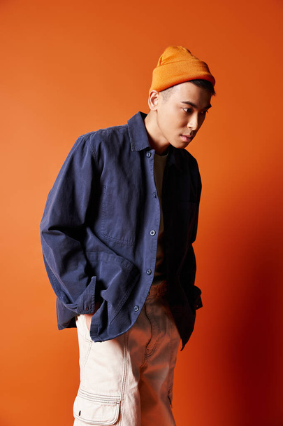 Стильный молодой азиат в голубой рубашке и оранжевой шляпе уверенно позирует на ярком оранжевом фоне. - Фото, изображение