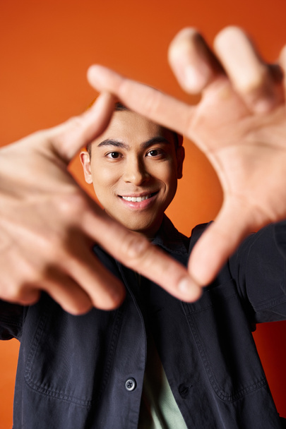 スタイリッシュな服装のハンサムなアジア人男性は,オレンジ色のスタジオ背景に対して彼の手でハートを形作ります. - 写真・画像