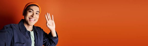 Een stijlvolle Aziatische man in een blauw jasje gebaart met zijn hand tegen een levendige oranje achtergrond in een studio setting. - Foto, afbeelding