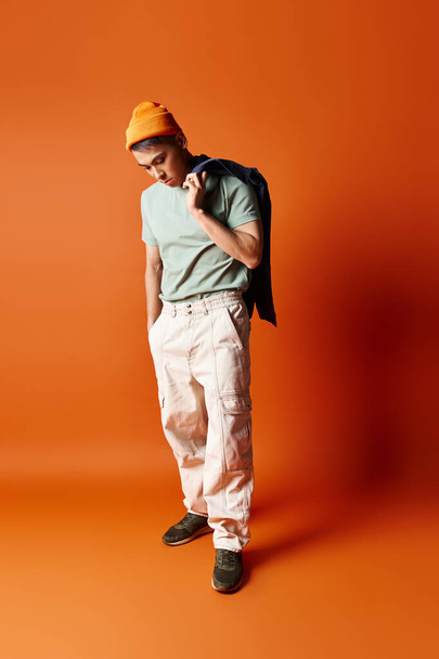 Ein gut aussehender asiatischer Mann posiert selbstbewusst in grünem Hemd und weißer Hose vor orangefarbenem Hintergrund in einem Studio-Setting. - Foto, Bild