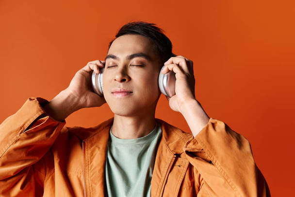 Ein hübscher asiatischer Mann in stylischer Kleidung mit Kopfhörern an den Ohren auf orangefarbenem Hintergrund. - Foto, Bild