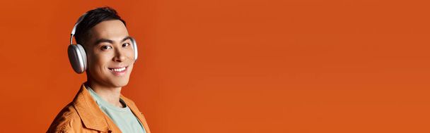 Красивый азиат в стильной одежде в наушниках и пиджаке, снятый на фоне оранжевой студии. - Фото, изображение