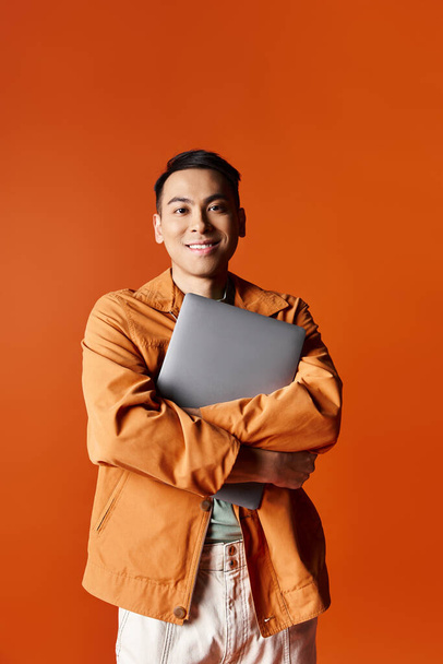 Um homem asiático elegante com braços cruzados, segurando com confiança um laptop contra um pano de fundo laranja. - Foto, Imagem