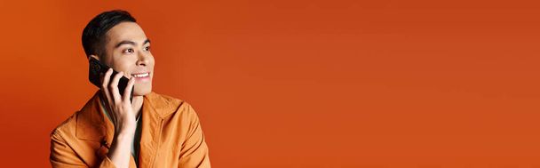 Ein stilvoller asiatischer Mann unterhält sich auf einem Handy vor einem grellen orangefarbenen Hintergrund. - Foto, Bild