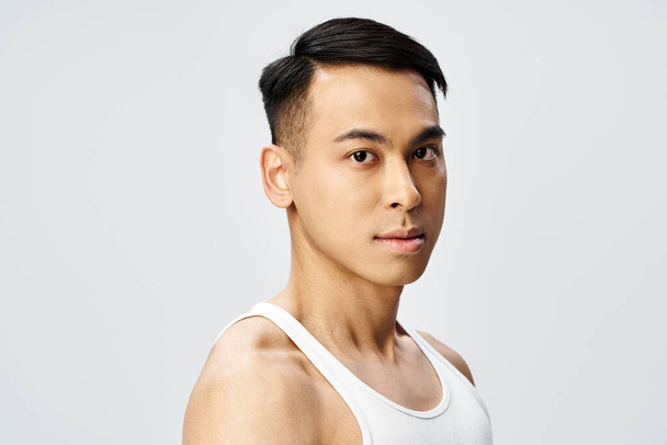 Un hombre asiático guapo en una camiseta sin mangas da una pose segura en un estudio gris. - Foto, imagen
