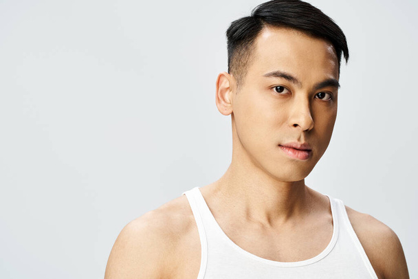 A young Asian man in a tank top strikes a pose in a grey studio setting, exuding confidence and tranquility. - Photo, Image