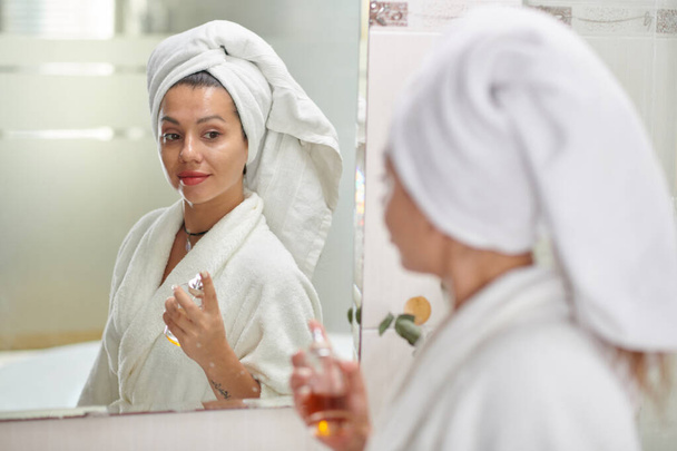 Jeune femme avec serviette sur la tête debout devant le miroir dans la salle de bain et pulvérisation de parfum de bouteille sur sa peau après la douche - Photo, image
