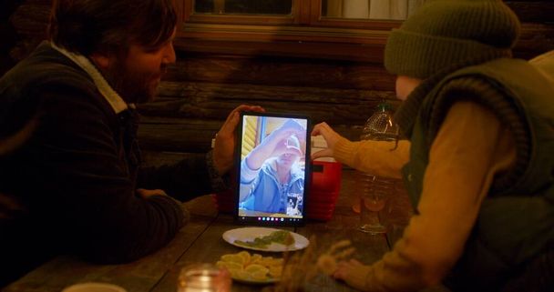 Homem adulto detém tablet computador enquanto sentado ao ar livre à noite durante o jantar com as crianças e conversando com a mulher por videochamada. Família turística descansar juntos durante as férias no campo. - Foto, Imagem