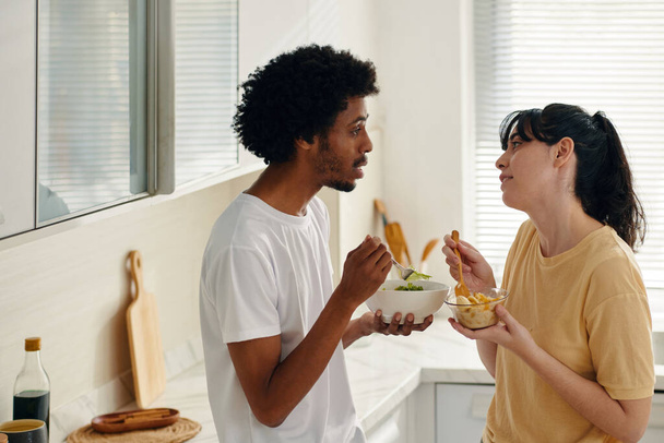 Πλευρική άποψη του νεαρού πολυπολιτισμικού ζευγαριού με σαλάτα σε μπολ τρώγοντας πρωινό και κοιτάζοντας ο ένας τον άλλο στην κουζίνα - Φωτογραφία, εικόνα