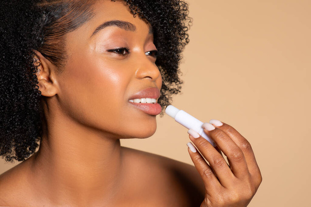 Profiel van jonge zwarte vrouw met krullend haar van toepassing witte lip balsem, gericht op lip zorg tegen zachte beige studio achtergrond, benadrukken schoonheid - Foto, afbeelding